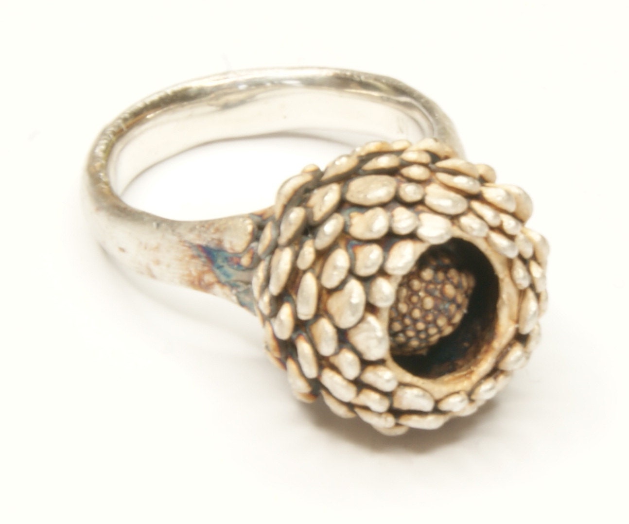 Handmade silver ring with hand-formed pinecone-like scales on the outer surface and sculptural granular interior, luminous white finish, worn on finger against aged mossy wood background.